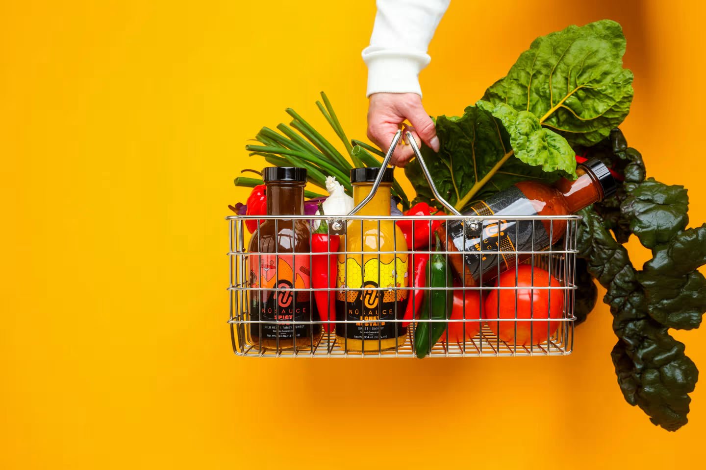 Hand holding a metal basket filled with groceries against a yellow background