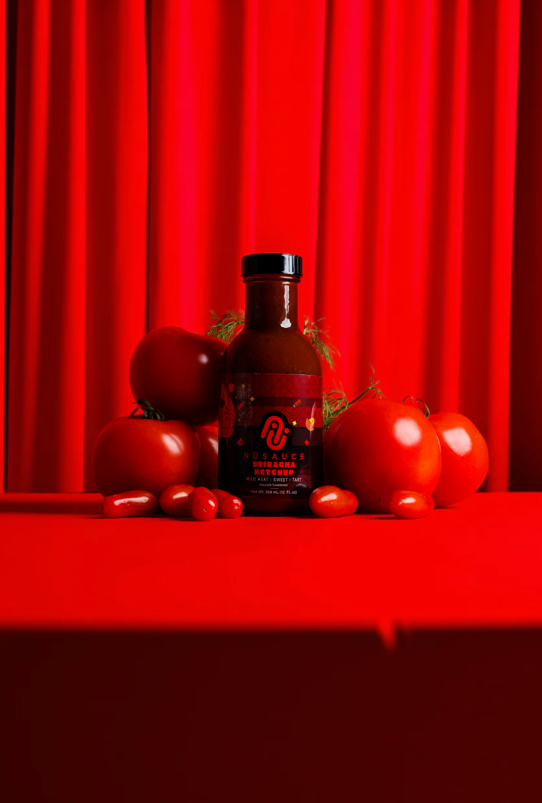Bottle of hot sauce surrounded by tomatoes against a red curtain background